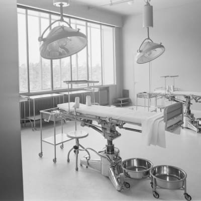 1940-luvun sairaalan toimenpidehuone, jossa on kaksi sänkyä, lamppuja ja muita sairaanhoidollista välineistöä siististi paikoillaan.
