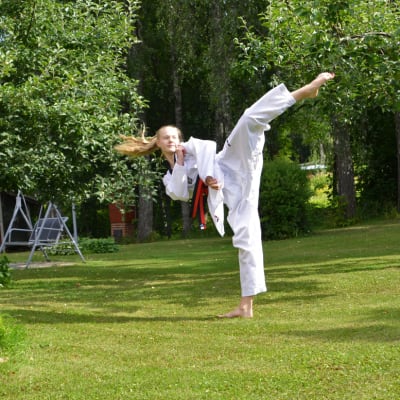En ung kvinna i taekwondodräkt sparkar upp i luften. Hon står på en gräsmatta framför några äppelträd.