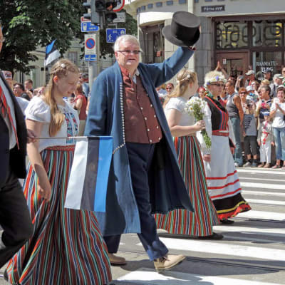 Kansallispukuinen Savisaar heiluttaa hattuaan. Vierssä kansallispukuisia naisia, kadun varressa ihmiset taputtavat.