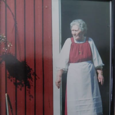 80-vuotias rouva kansallispuvussa punaisen tuvan portailla aurinkoisena kesäpäivänä.