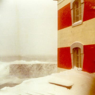Vinterstorm vid randig fyr, havet sköljer över mur, snö är samlad under fönstret till en rödvitrandig fyr.