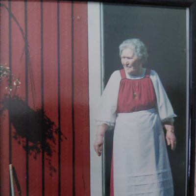 80-vuotias rouva kansallispuvussa punaisen tuvan portailla aurinkoisena kesäpäivänä.
