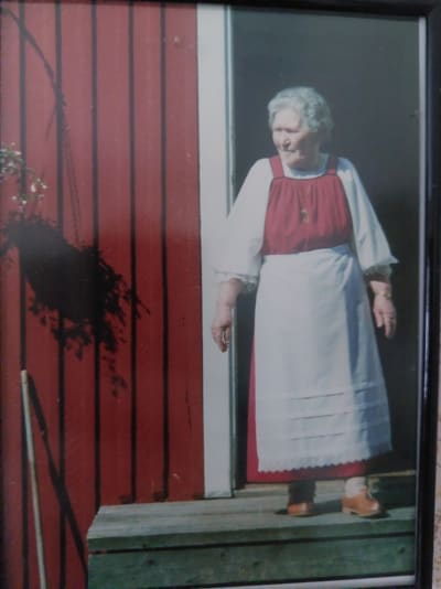 80-vuotias rouva kansallispuvussa punaisen tuvan portailla aurinkoisena kesäpäivänä.