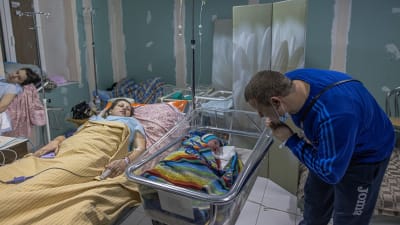 En kvinna ligger i en sjukhussäng. Bredvid henne ligger en nyfödd bebis i en liten säng. En man talar med dem.
