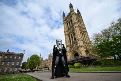 A man in some kind of superhero costume in black and gray, with a black cape and a dustbin for a head stands and gestures in front of Westminster Abbey.