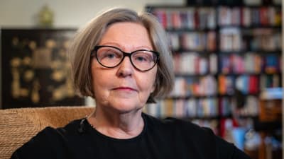 Anja Lahtinen hemma, i svart klänning med bokhylla i bakgrunden.
