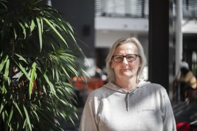 En kvinna ser bestämt in i kameran. Bredvid henne står en palm. I bakgrunden syns en skolmatsal.