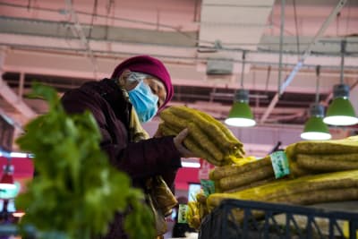 Kvinna i munskydd kollar på grönsaker i affär.