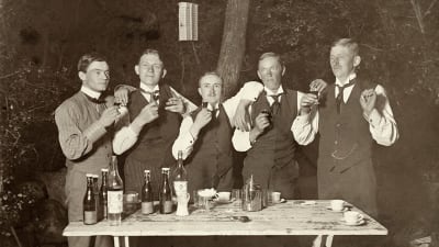 Fem män höjer sina snapsglas ute i en trädgård stående vid ett trädgårdsbord med kaffekoppar och flaskor