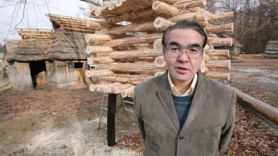 En medelålders man står framför träbyggnader med händerna bakom ryggen.