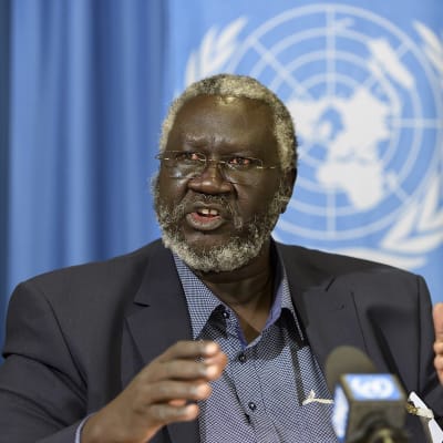 Harmaa hiuksinen, silmälasipäinen mies tummassa puhuu mikrofonin edessä, taustalla sininen verho, jossa näkyy YK:n logo. 