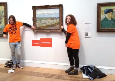 Två aktivister i orange t-skjortor står med varsin hand fastlimmad i ramen till Vincent van Goghs målning Blommande persikaträd.