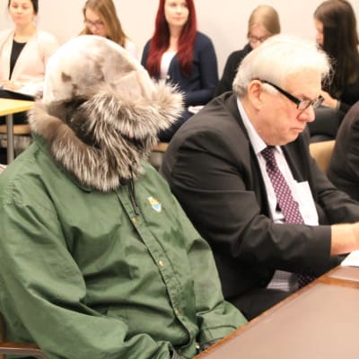 Pekka Seppänen sitter i rättssalen med en grön jacka och huvan framför ansiktet.