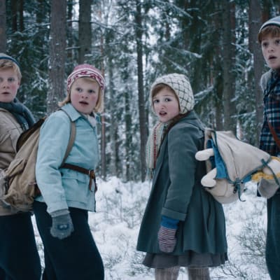 Fyra skrämda barn står i ett skogsbryn, vänder sig om i hast och tittar snett förbi kameran.