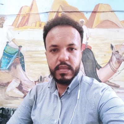 Mohamed är en ung man i 30-års åldern. Han har på sig en kragskjorta. Bakom honom är en väggmålning med kameler.