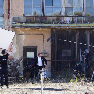 I en filminspelning springer en man i vit skjorta och kavaj ut ur ett hus. En man står med en stor solreflektor, en annan med en filmkamera och en tredje med en mikrofon på en lång stång  kring honom.