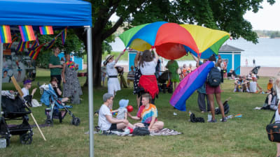 Många människor sitter på en gräsmatta vid en strand. Några håller upp en regnbågsflagga.