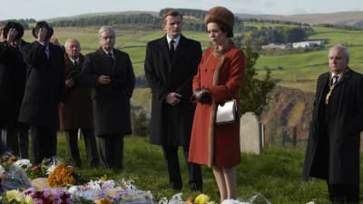 Storbritanniens drottning klädd i en röd kappa står på en begravningsplats i ett naturskönt landskap omgiven av svartklädda män.