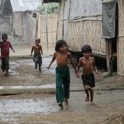 Lapsia juoksee sateessa. Bamburakenteisia taloja kylän raitilla. 