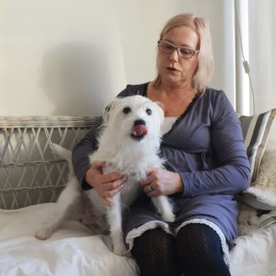 En kvinna sitter i en soffa och håller om en liten vit hund. Kvinnan tittar på hunden som slickar sig om munnen.