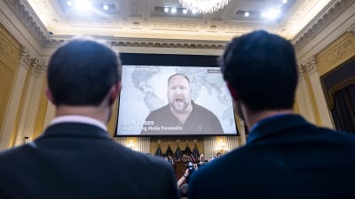 Alex Jones visas på en storskärm i en sal med människor. Under skärmen syns amerikanska flaggor.
