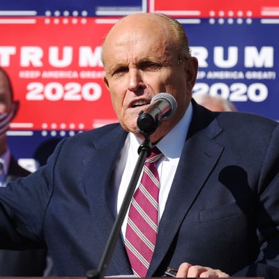 Man i blå kostym och rödrandig slips står framför vägg där det står Trump 2020. 