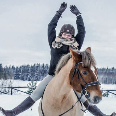 En ung kvinna sitter på en häst. Hon sträcker upp händerna i luften och fötterna åt sidorna.