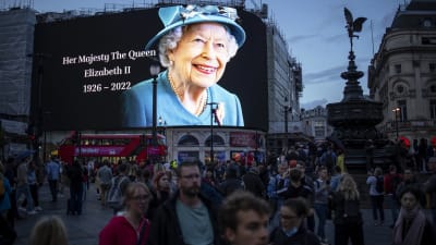 En folkmassa vid Piccadilly Circus i London nedanför en stor ljustavla med en bild av drottning Elizabeth II i blå hatt och dräkt med årtalen 1926-2022.