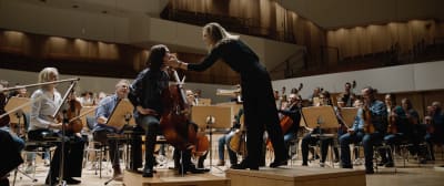 En kvinnlig dirigent stryker en kvinnlig cellist över kinden medan resten av orkestern tittar på.