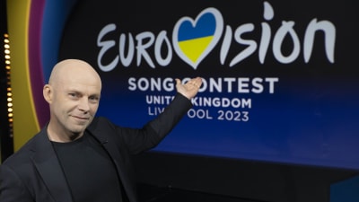 En skallig man i mörk kavaj står i en studio, ser in i kameran och pekar på logon för Eurovision Song Contest 2023.