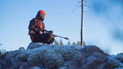 Istuva metsästäjä huurteisessa maisemassa, jossa näkyy lisäksi taivasta vasten kukkula ja yksi ohuehko puunrunko.