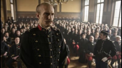 En gråhårig Alfred Dreyfus står i en domstolssal iklädd militäruniform, med en stor publik bakom sig. 