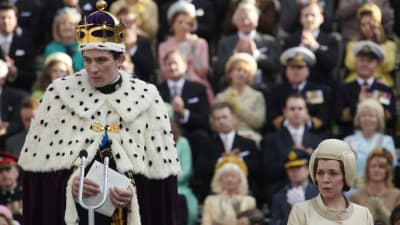 En ung man i hermelinpäls och en gyllne krona står på ett podium framför en mikrofon. I bakgrunden syns en festklädd publik. 