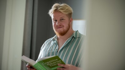 Sami-Petteri Asikainen pitelee kädessään vihreää oppikirjaa, jonka kannessa lukee Cognitive Behavior Therapy. Hän katsoo kameraan hymyillen.