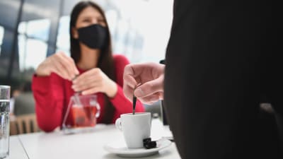 Kvinna i munskydd och man som bara syns bakifrån sitter på café