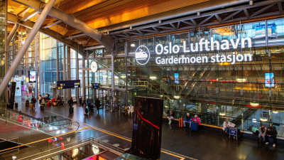 Flygplats där människor står och väntar med resväskor. På en vägg står det "Oslo Lufthavn".