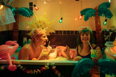 En ung kille och en ung tjej i ett badkar med uppblåsbara palmer i bakgrunden och drinkar i händerna.