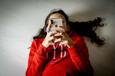 En flicka ligger på golvet och scrollar i sin telefon. Telefonen täcker hennes ansikte.