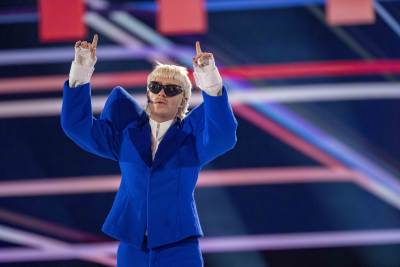 En blond man iklädd solglasögon och klarblå scenkostym står på en scen med händerna höjda över huvudet.