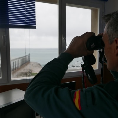 Vartiotorinissa oleva rajapoliisi katsoo kiikareille merelle, ulkona näkyy teräsaita, rantaa ja merta.