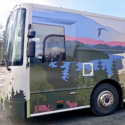 Metsäinen maalattu maisema kirjastoauton kyljessä. Auto seisoo kirjaston pihalla.