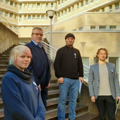 Neljä henkilöä poseeraa päivänvalossa rakennuksen portailla ja katsoo kameraan