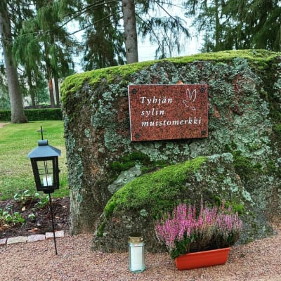 Sammaleinen kivimuistomerkki, jossa on kivinen laatta tekstillä "Tyhjän sylin muistomerkki". Laatassa on myös linnun kuva. Kiven edellä on kaksi kynttilälyhtyä ja kanervaistutus.
