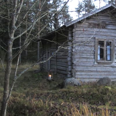 Harmaan, vanhan rakennuksen vieressä palaa kynttilä. Talo on Laitilan Kaariaisten kylän mäkitupa.