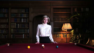 Kristen Stewart i rollen som prinsessan Diana i filmen Spencer. Hon står i ett mörkt rum med träpanel framför ett biljardbord. Ho när iklädd en vit blus och mörk underdel och har en allvarlig uppsyn.
