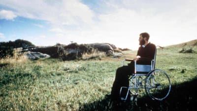 Parrakas mies tummissa vaatteissa istuu pyörätuolissa katsellen kallioista ja ruohikkoista nummimaisemaa.