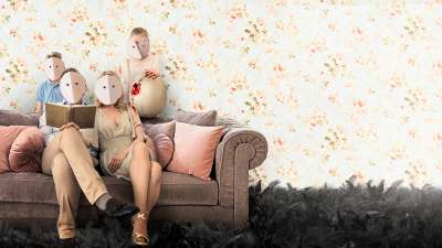 Perhe istuu sohvalla kukkatapetoidun seinän edessä, kaikkien kasvoja peittävät valkeat pahviset naamarit.