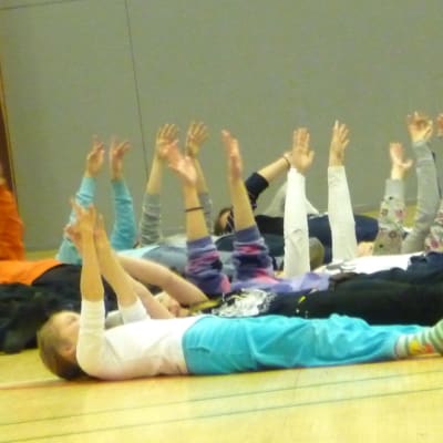 Lapsia makaa koulusalin lattialla kädet ylhäällä. 