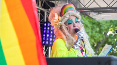 En person med stort hår och mycket smink talar i en mikrofon. Bredvid vajar en regnbågsflagga.
