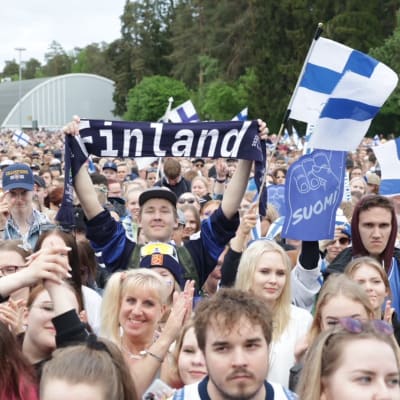 Yleisömerta hymyilemässä ja taputtamassa. Yleisön keskellä mies nostamassa huivia, jossa lukee "Finland".